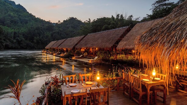 A riverside dining area with thatched-roof huts and wooden tables set for dining. - River Kwai Jungle Rafts (Sai Yok)