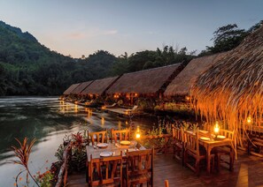 A riverside dining area with thatched-roof huts and wooden tables set for dining. - River Kwai Jungle Rafts (Sai Yok)