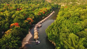 A long wooden boat docked along a river surrounded by dense forest. - River Kwai Jungle Rafts (Sai Yok)