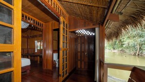 A wooden cabin with a thatched roof, a wooden floor, a bed, a toilet, and a window overlooking a river. - River Kwai Jungle Rafts (Sai Yok)