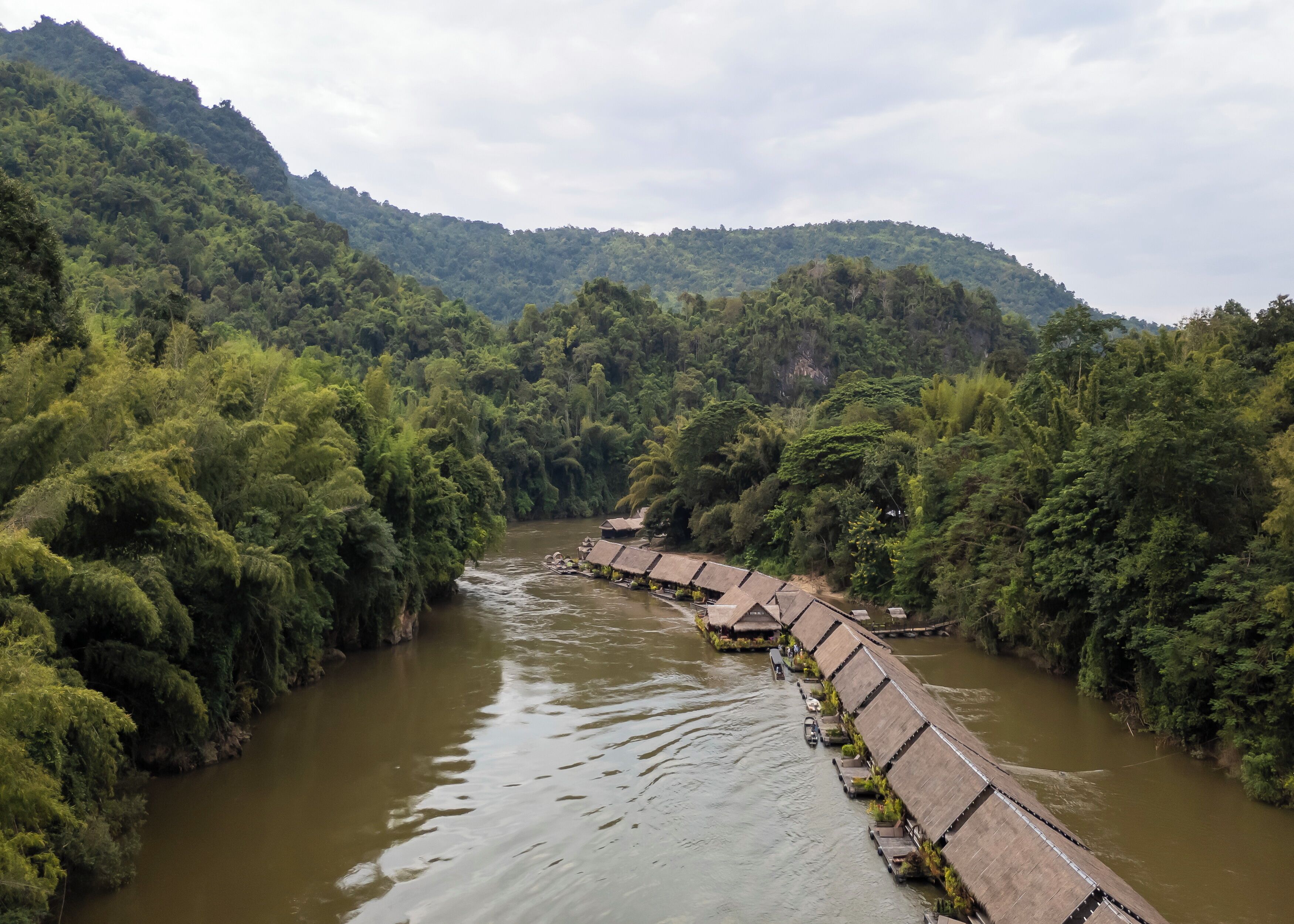 A river with a series of thatched-roof structures along its banks, surrounded by dense greenery and hills.