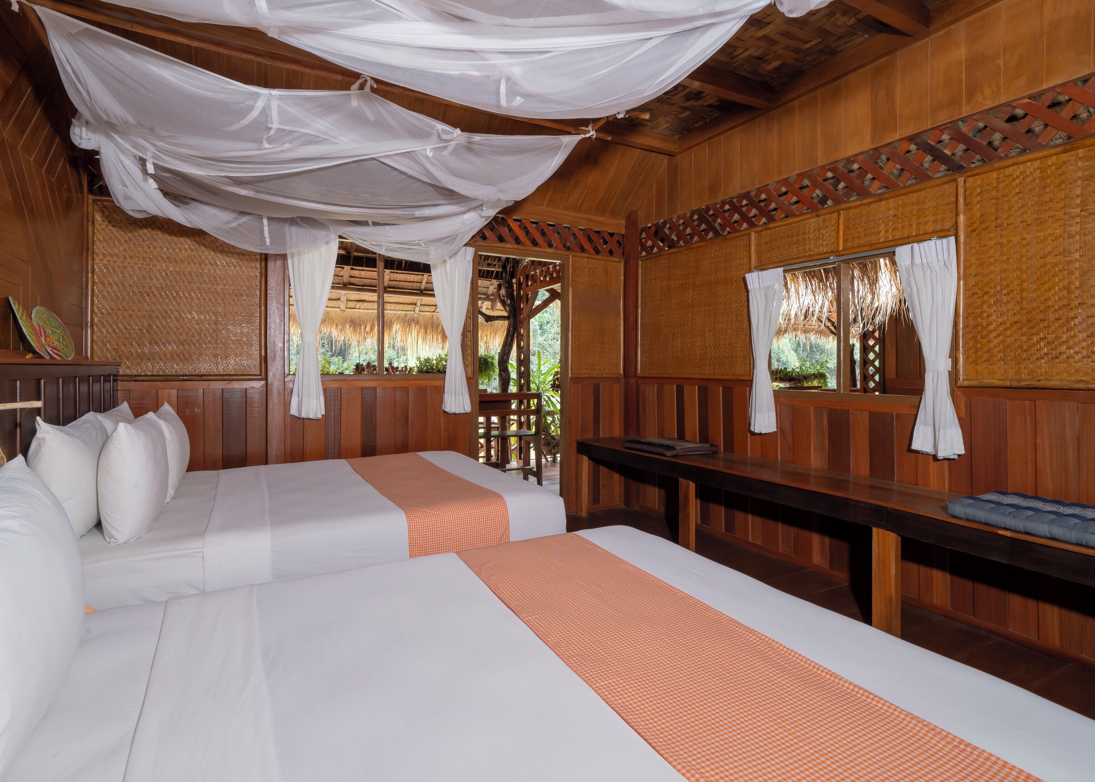 A room with two beds, wooden walls, a woven ceiling, and a view of greenery outside.