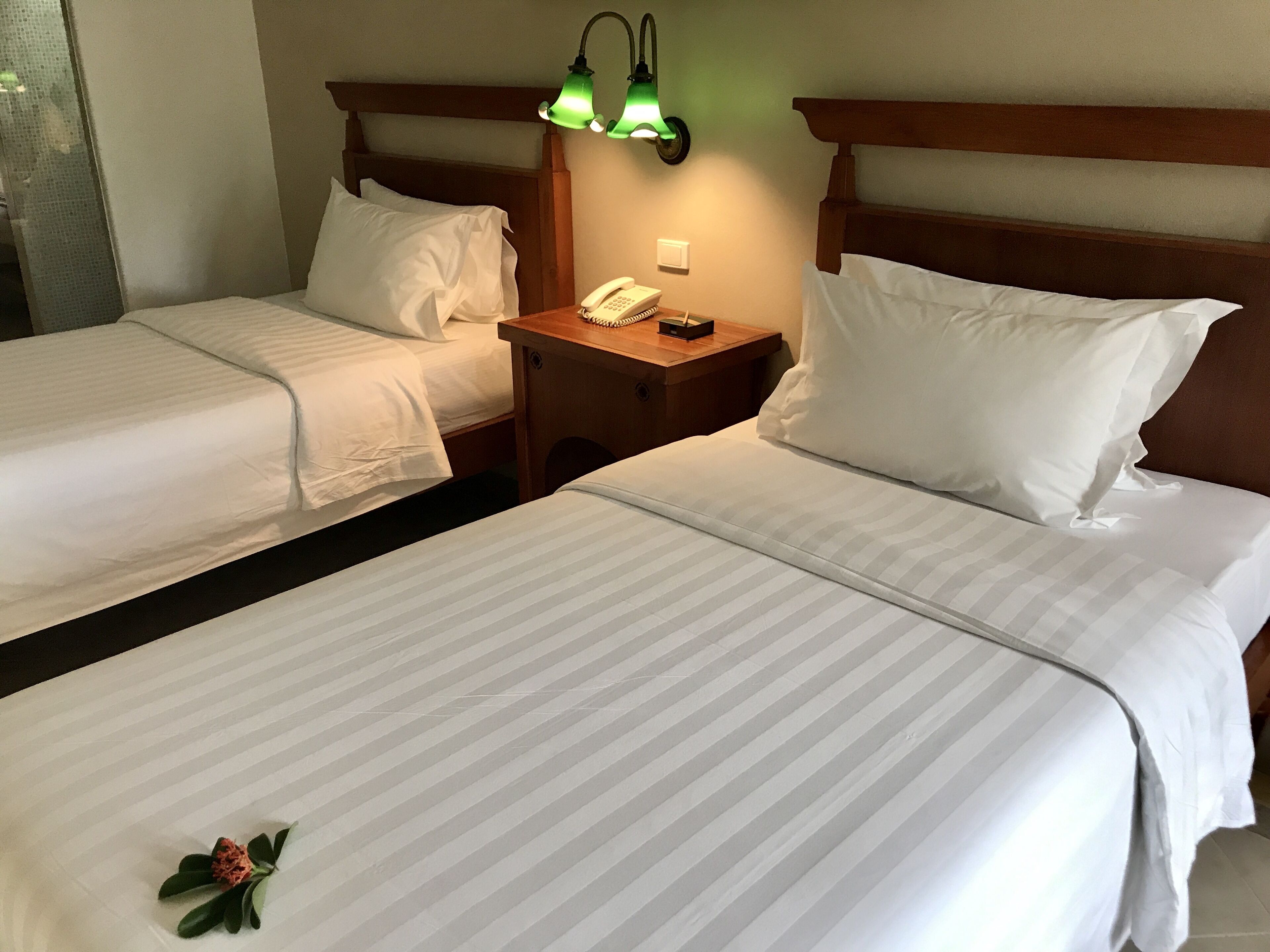 A hotel room with two single beds, each with white linens and a wooden headboard. There is a nightstand with a telephone and a green wall-mounted lamp between the beds.