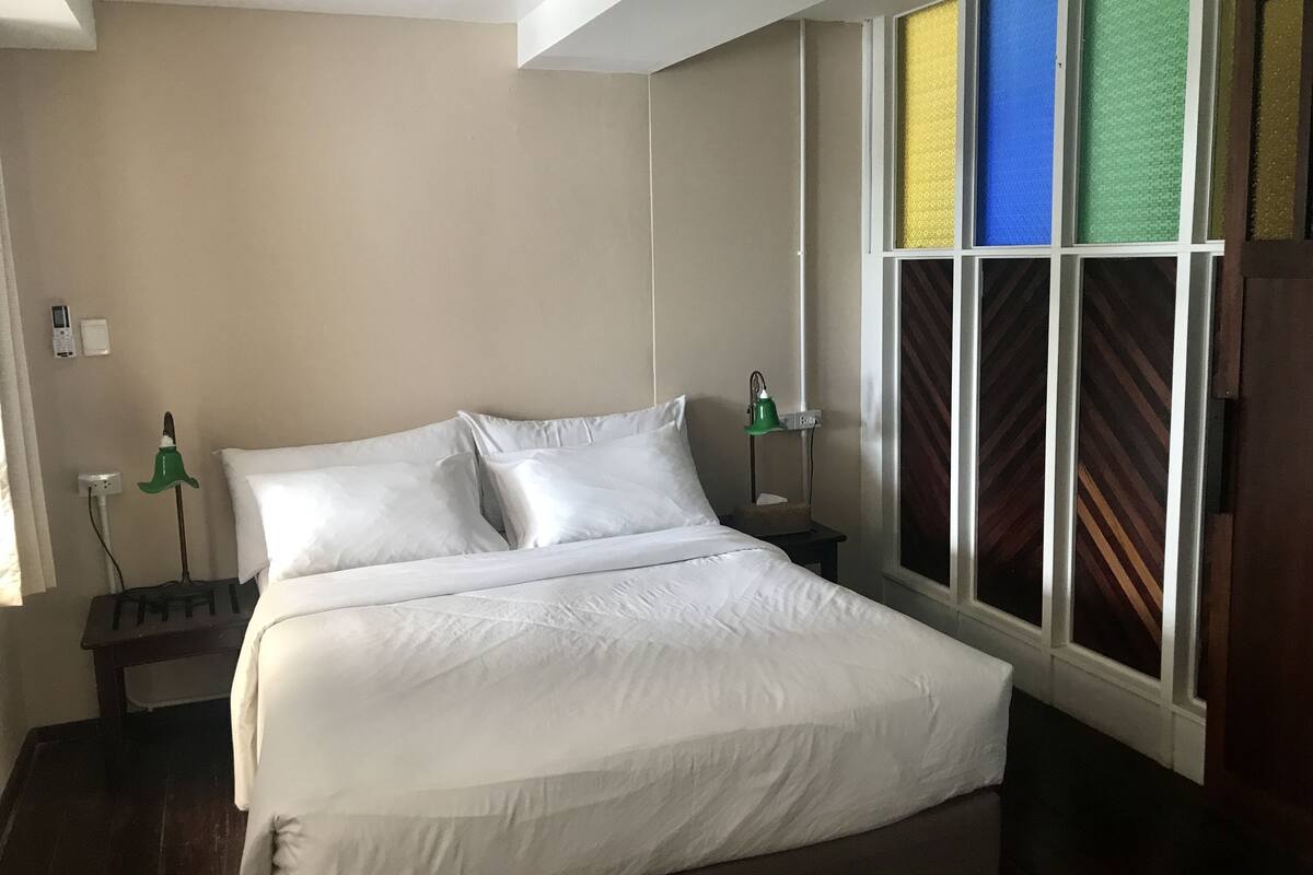 A bedroom with a bed, bedside lamps, a window with patterned curtains, and a small table.