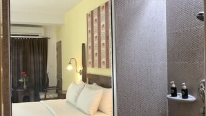 Une chambre d’hôtel dotée d’un grand miroir, d’un lit avec du linge de lit blanc, d’une table de chevet, d’un climatiseur fixé au mur et d’un espace douche avec un pommeau de douche à main.