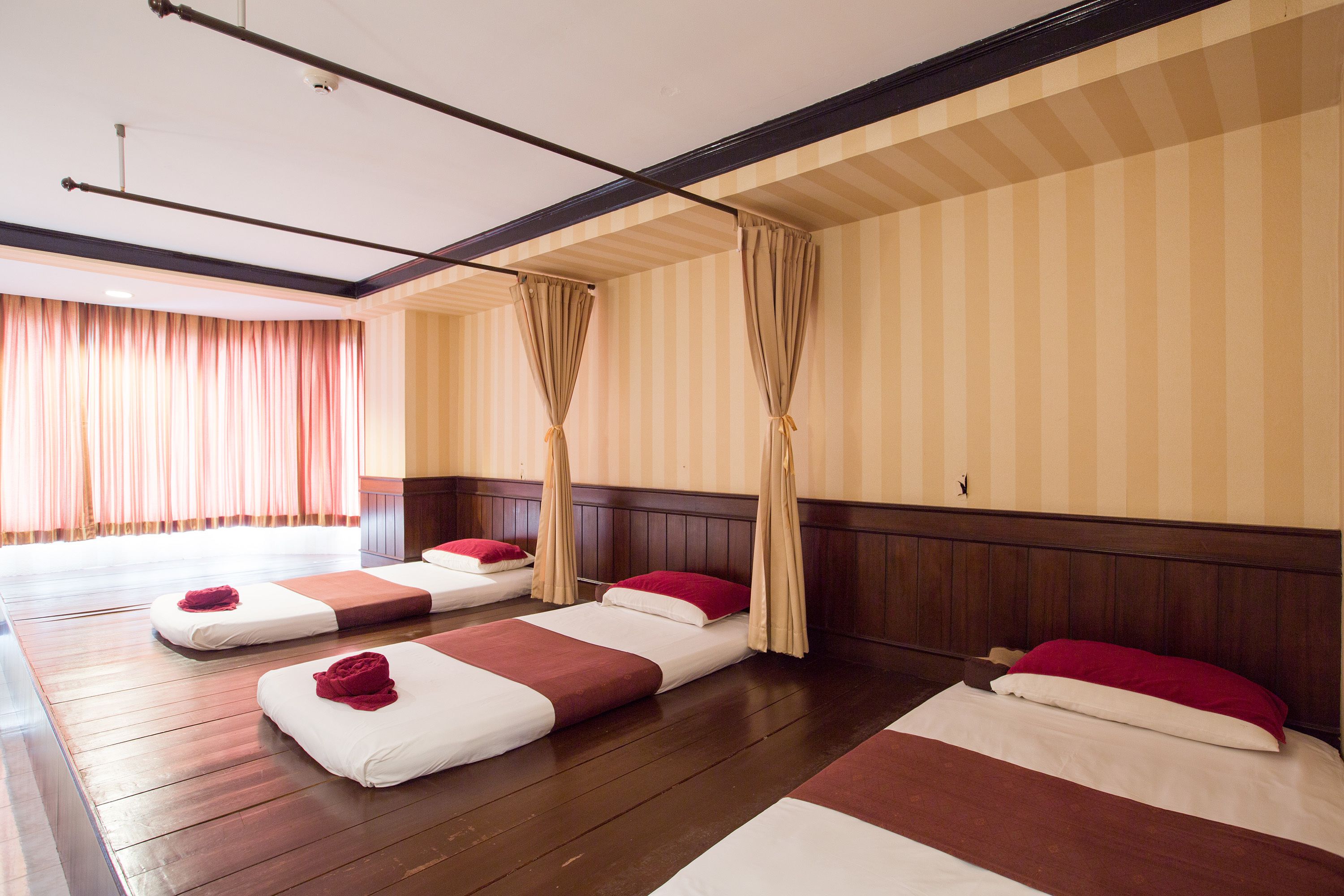 A hotel room with three single beds, wooden flooring, striped wallpaper, and a large window with curtains.