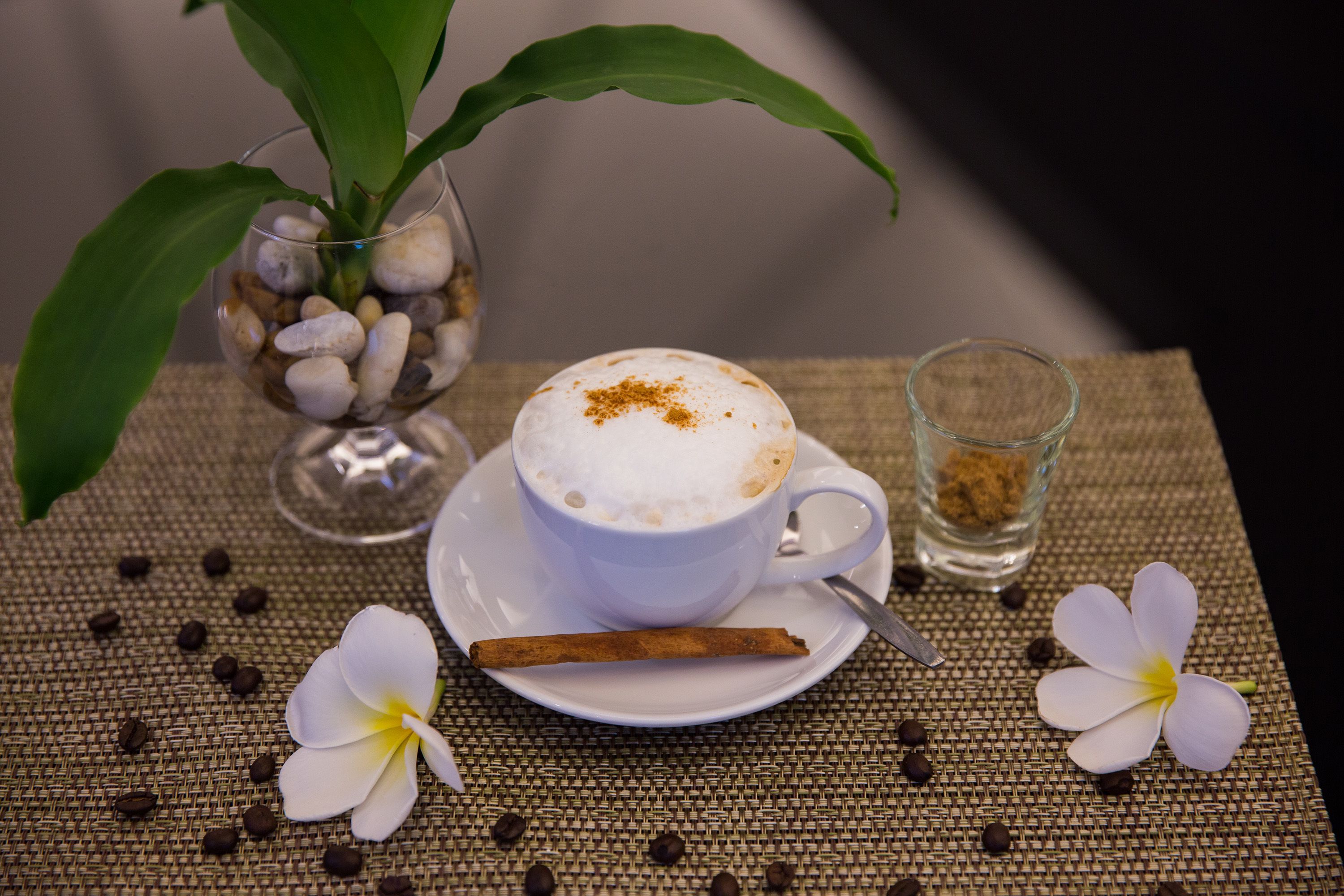 Una taza de café con una rama de canela sobre un mantel tejido, rodeada de granos de café y una flor.