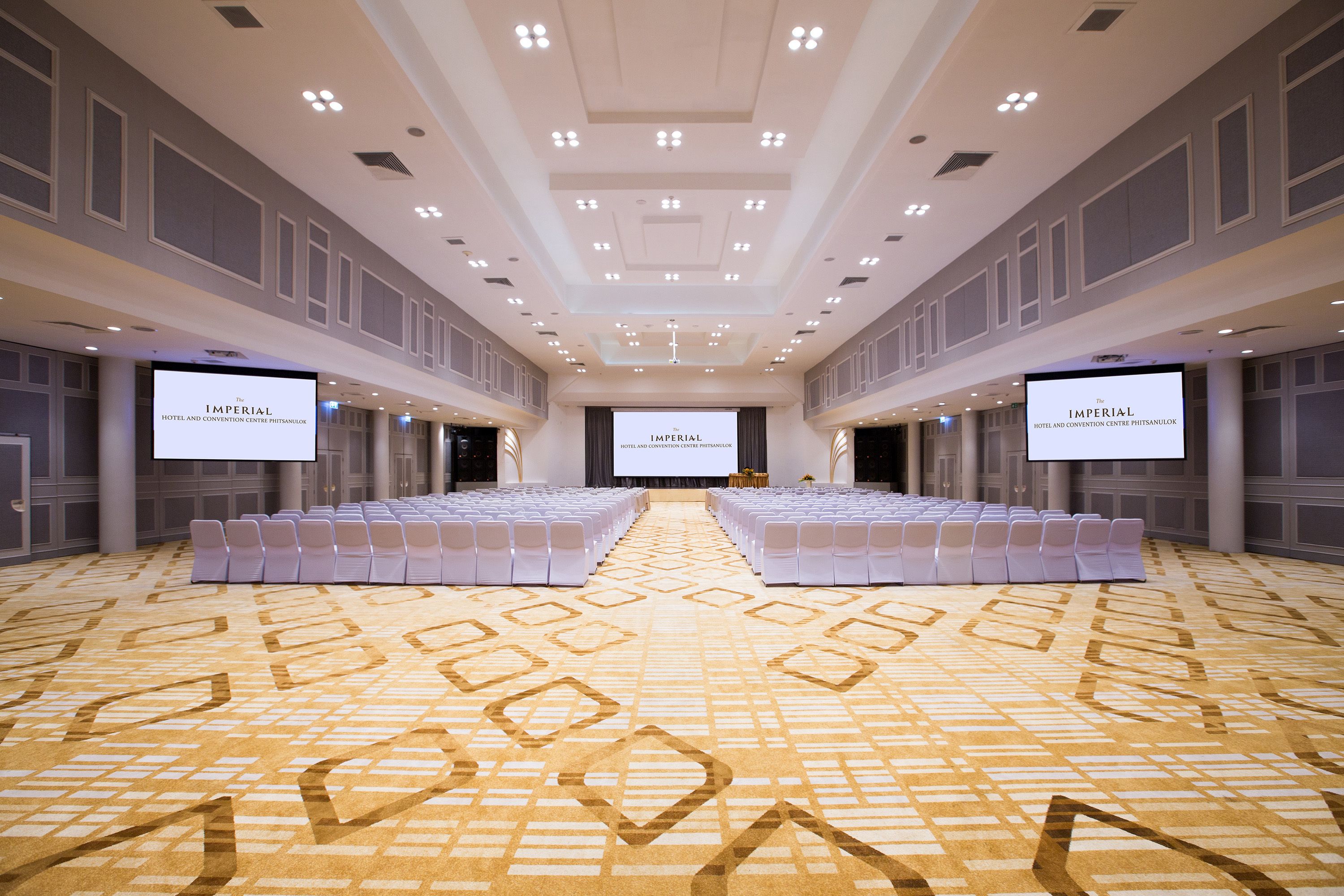 A large, well-lit conference room with a patterned carpet, rows of white chairs, and two large screens displaying the hotel name.