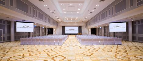A large, well-lit conference room with a patterned carpet, rows of white chairs, and two large screens displaying the hotel name.
