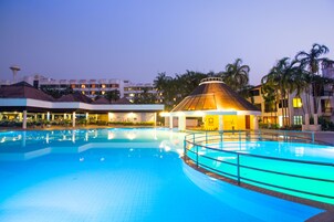 Una piscina con una estructura de techo de paja, rodeada de palmeras y edificios de hotel.