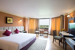 Habitación de hotel con una cama grande, un escritorio con televisor, un balcón con vistas y una zona de comedor con sillas.