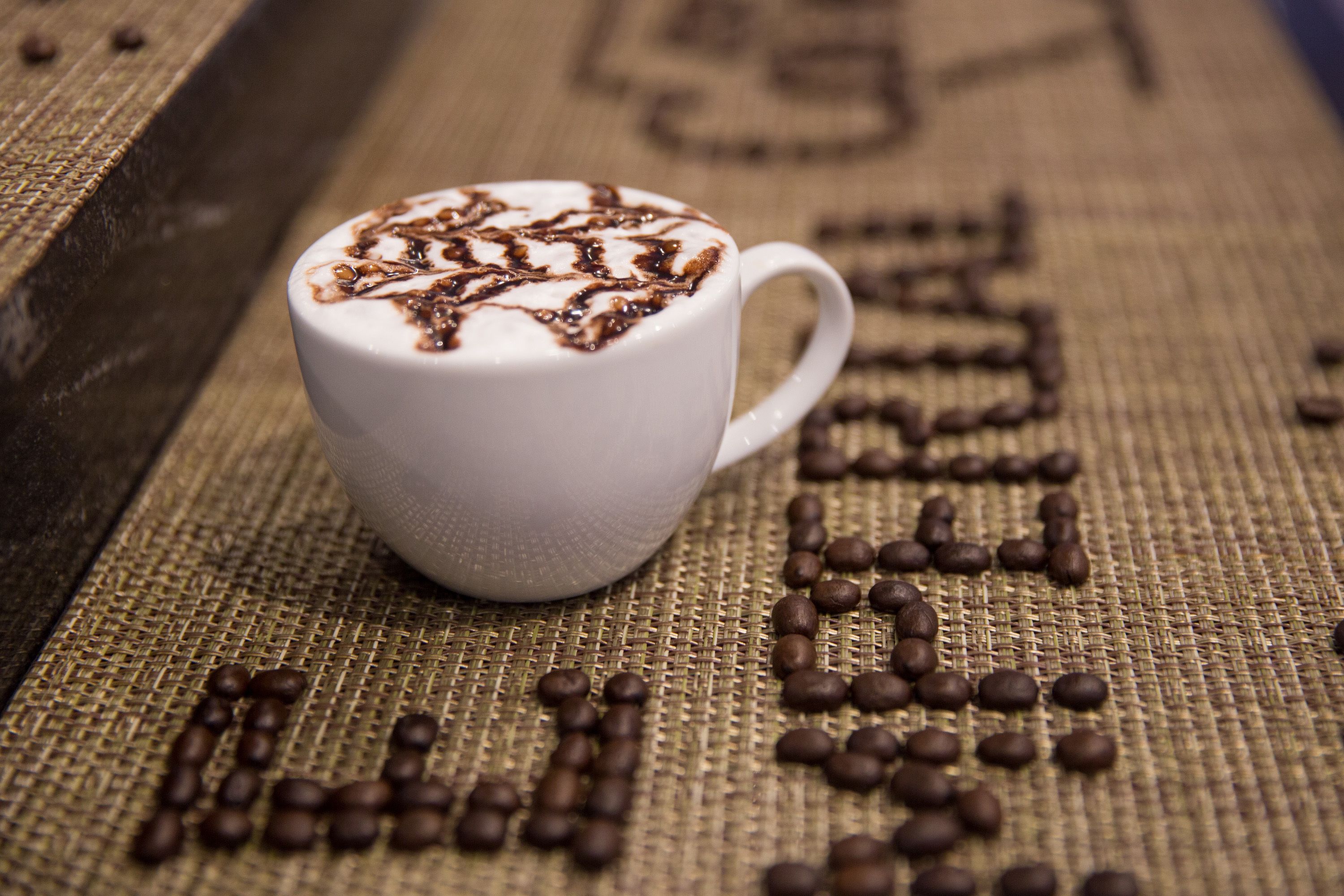 A white cup with a latte art design on a textured surface with coffee beans arranged in letters.