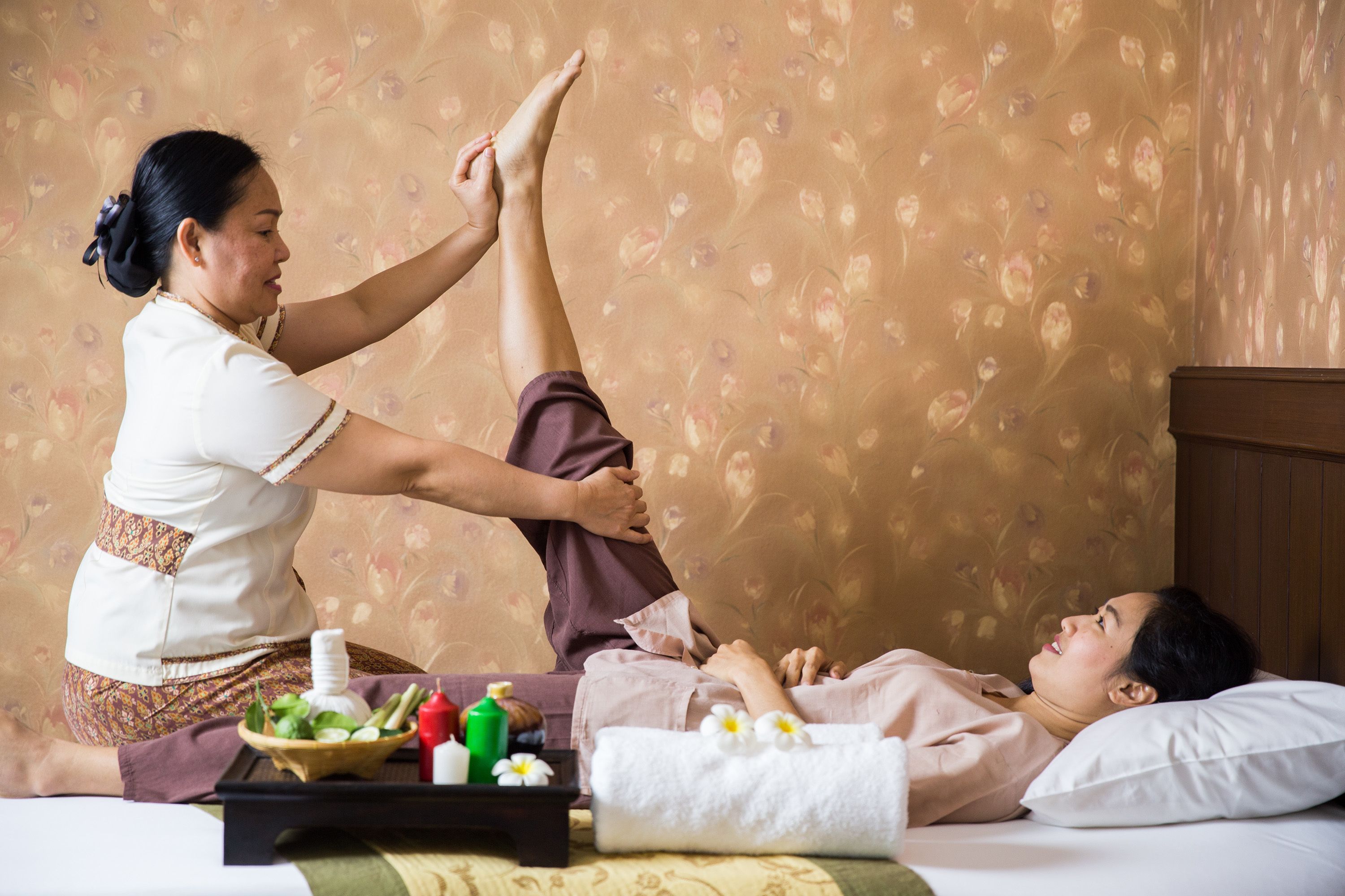 A person receiving a foot massage in a room with floral wallpaper, a tray with massage tools, and a bed with a white pillow.