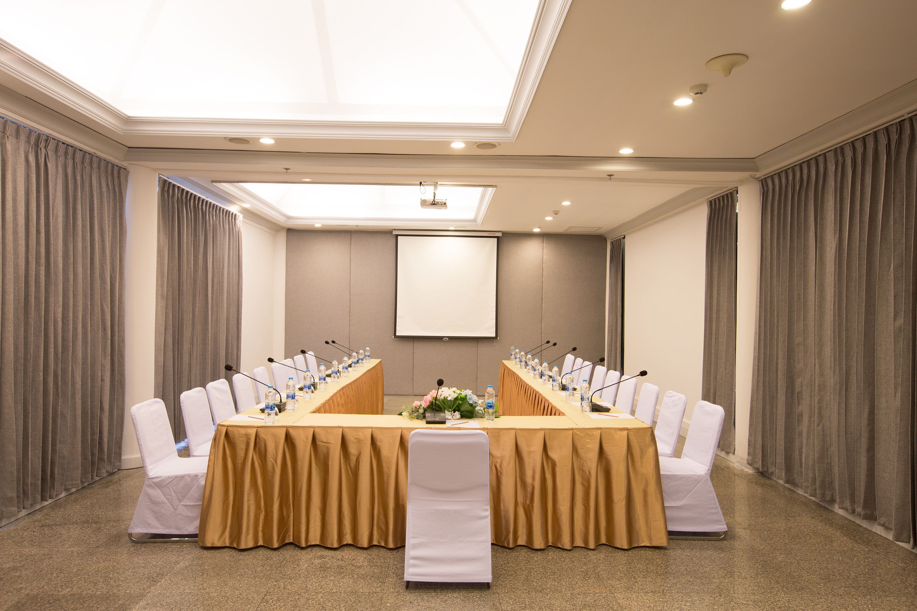 A conference room with a long table, white chairs, microphones, and a projector screen.
