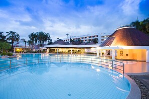 A swimming pool with a tiled deck, surrounded by palm trees and a building with a distinctive conical roof.