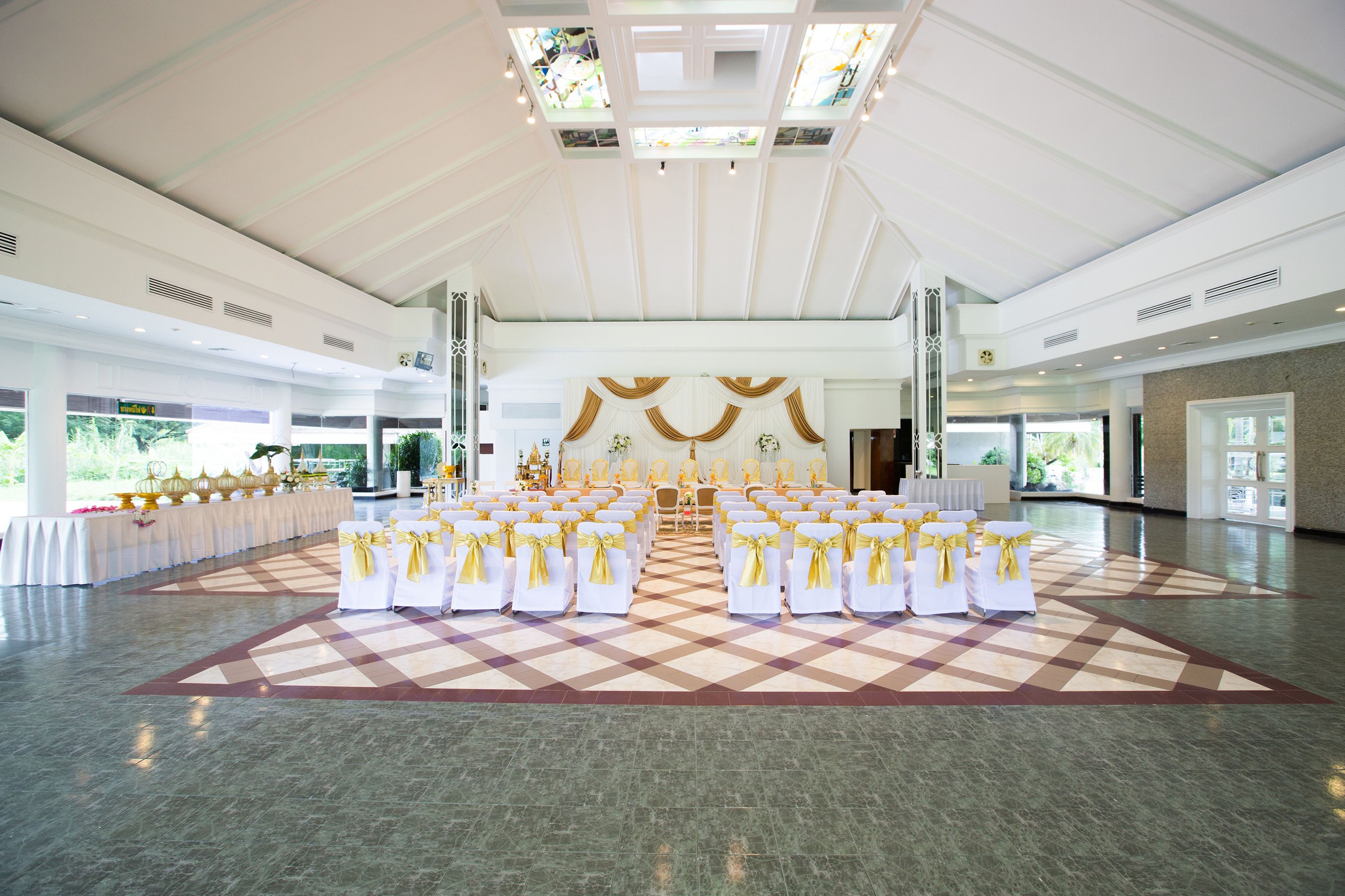 A spacious hall with a checkered floor, rows of white chairs with yellow sashes, and a central aisle leading to a stage with golden decorations.