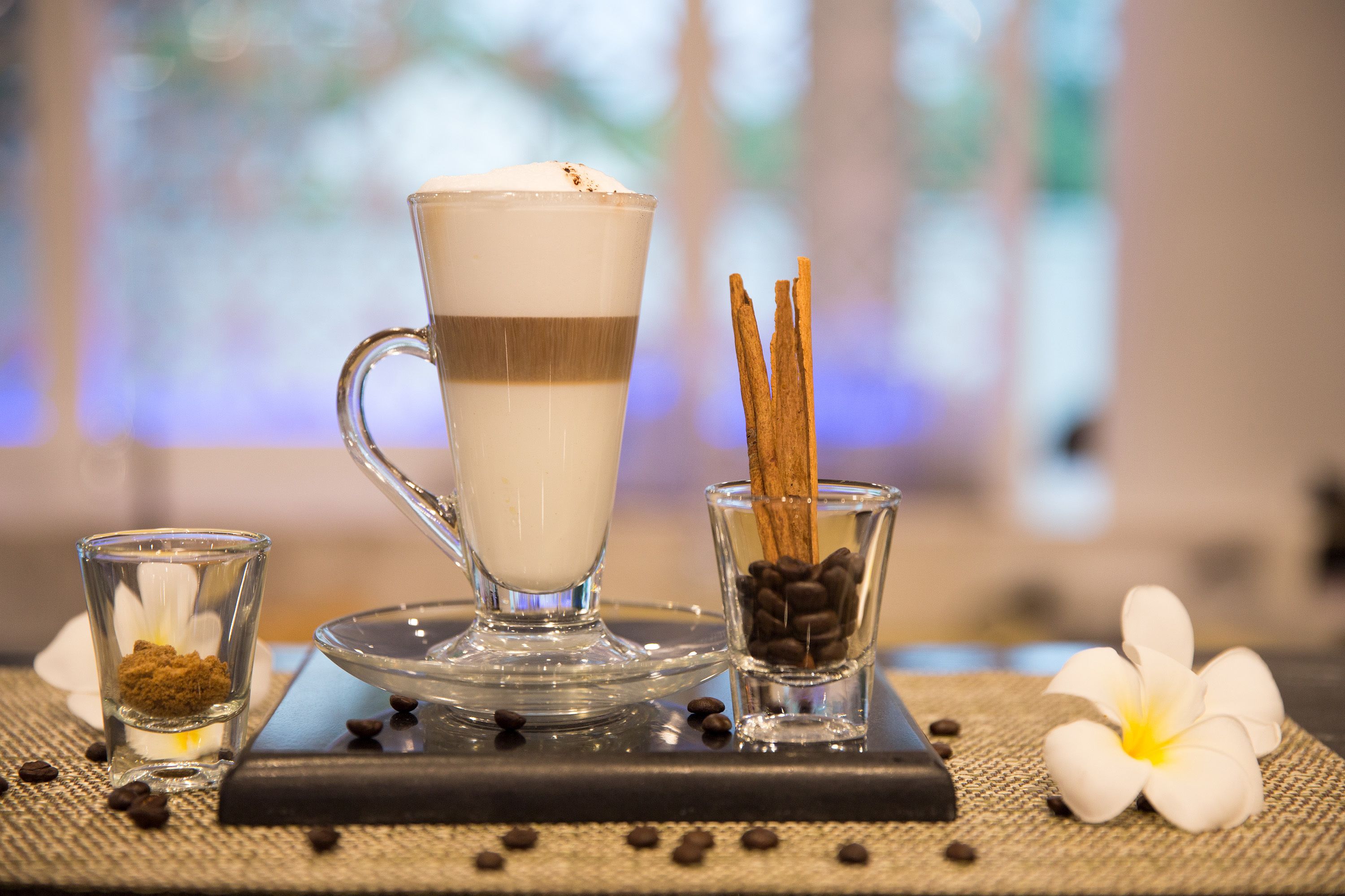 A clear glass cup with a layered beverage, a small glass with cinnamon sticks, and a flower on a textured surface.