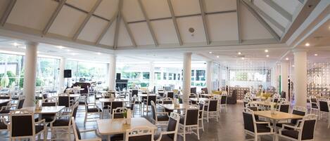 A spacious dining area with white tables and chairs, large windows, and a high ceiling.