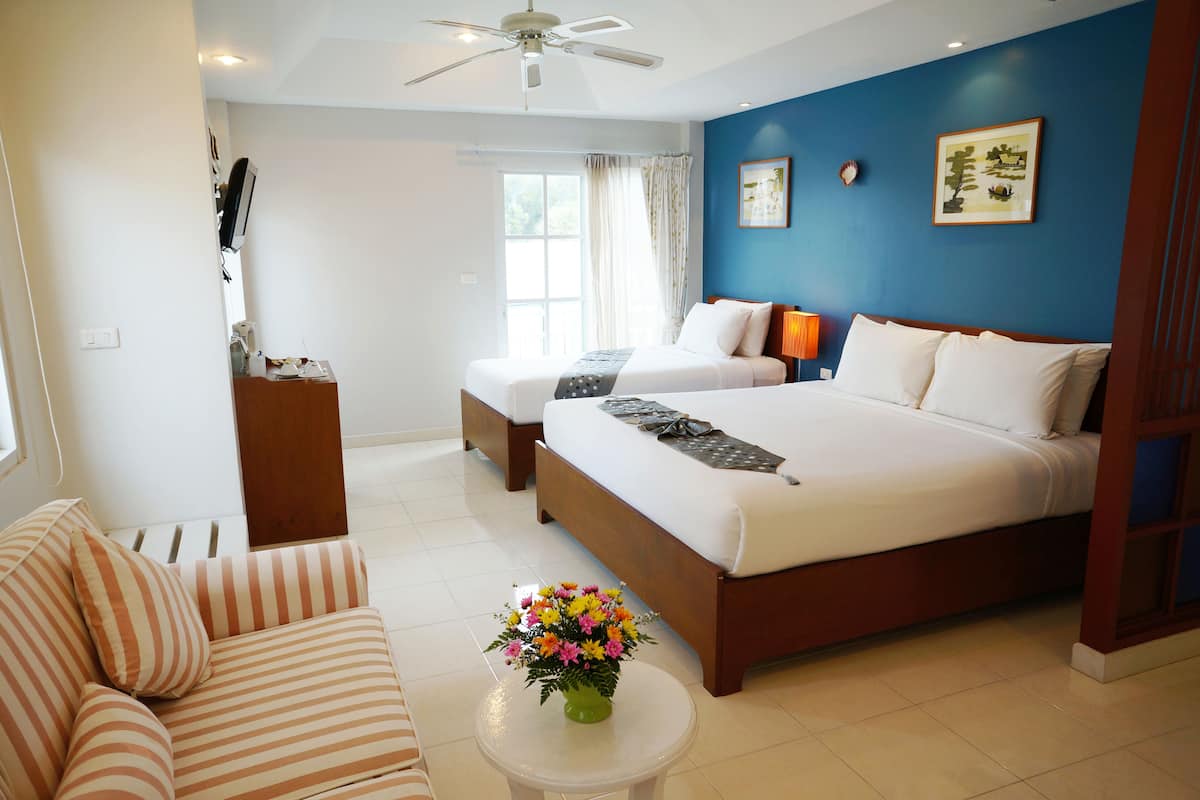 A hotel room with two beds, a sofa, a coffee table with flowers, a ceiling fan, and a window with curtains.