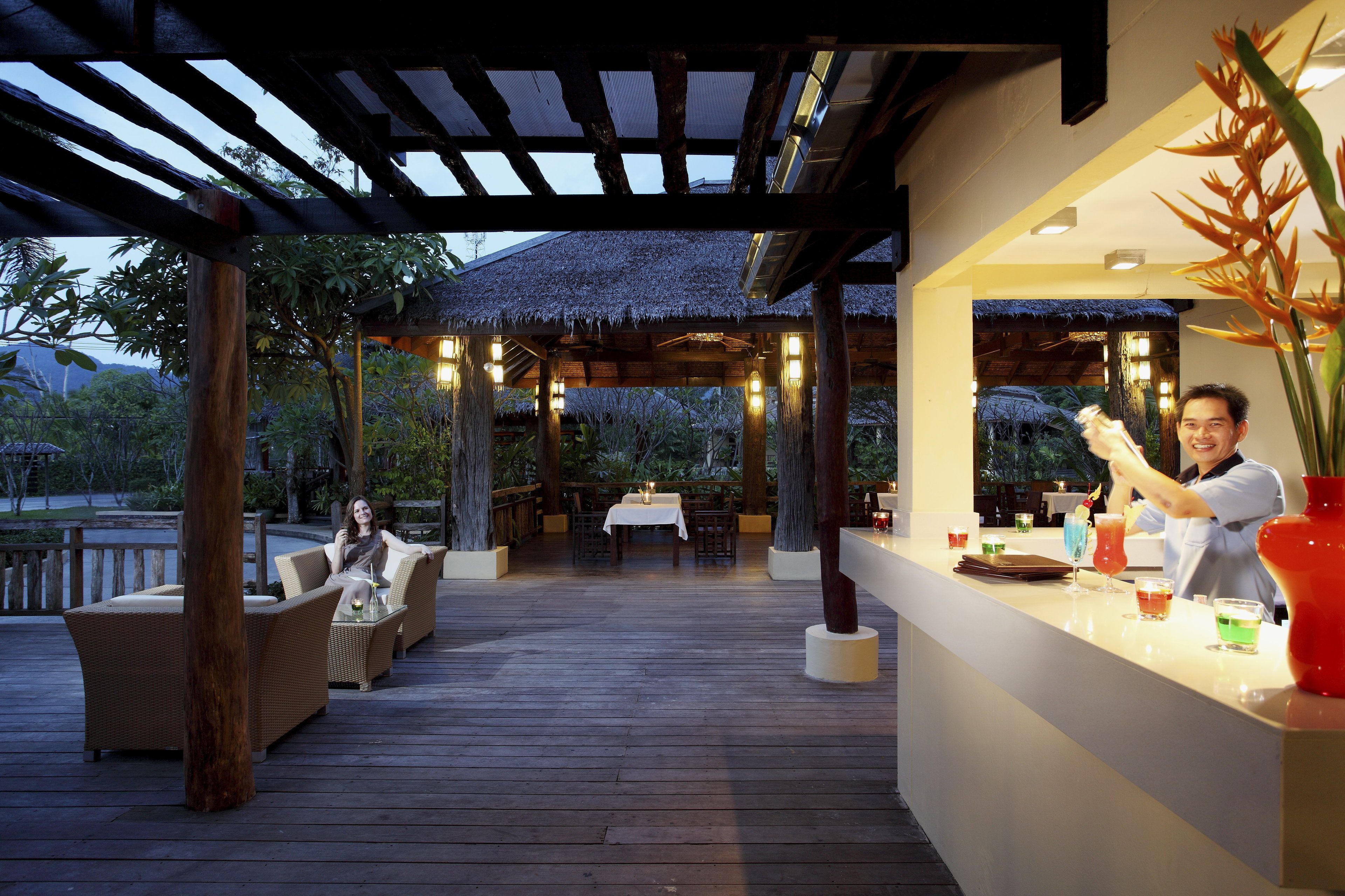 An outdoor dining area with wooden tables and chairs, a bar with a bartender, and a thatched-roof structure in the background.