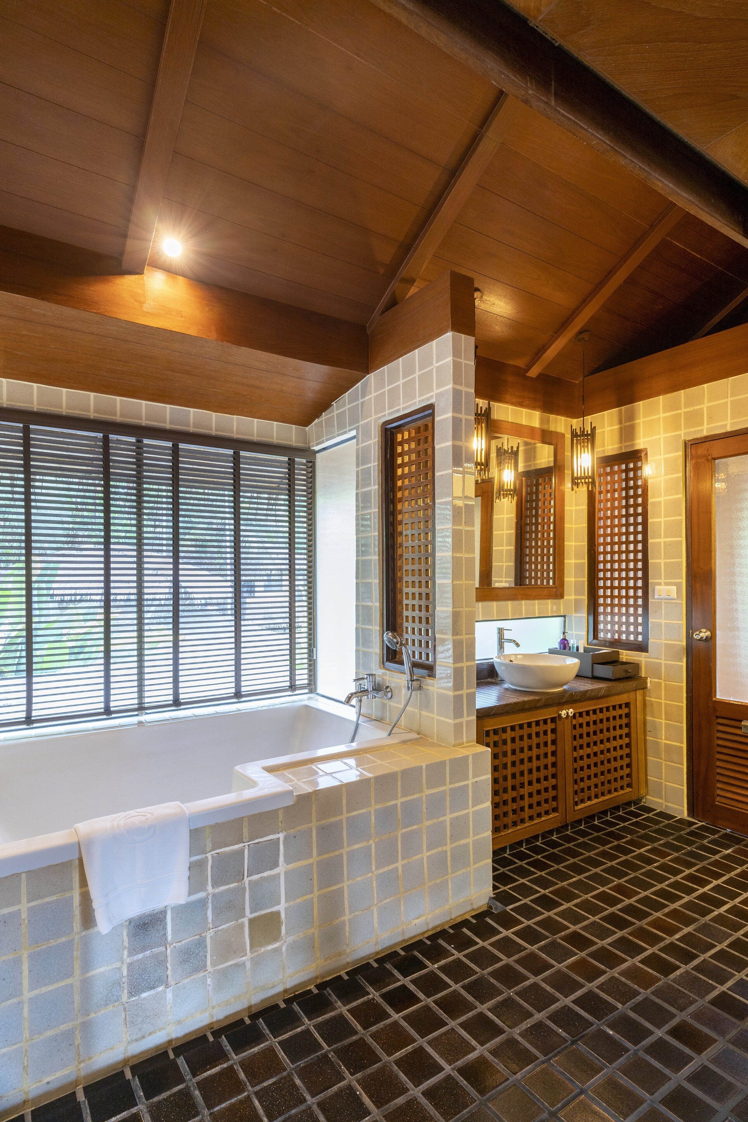 A modern bathroom with a large bathtub, a window with blinds, a sink, and a mirror.