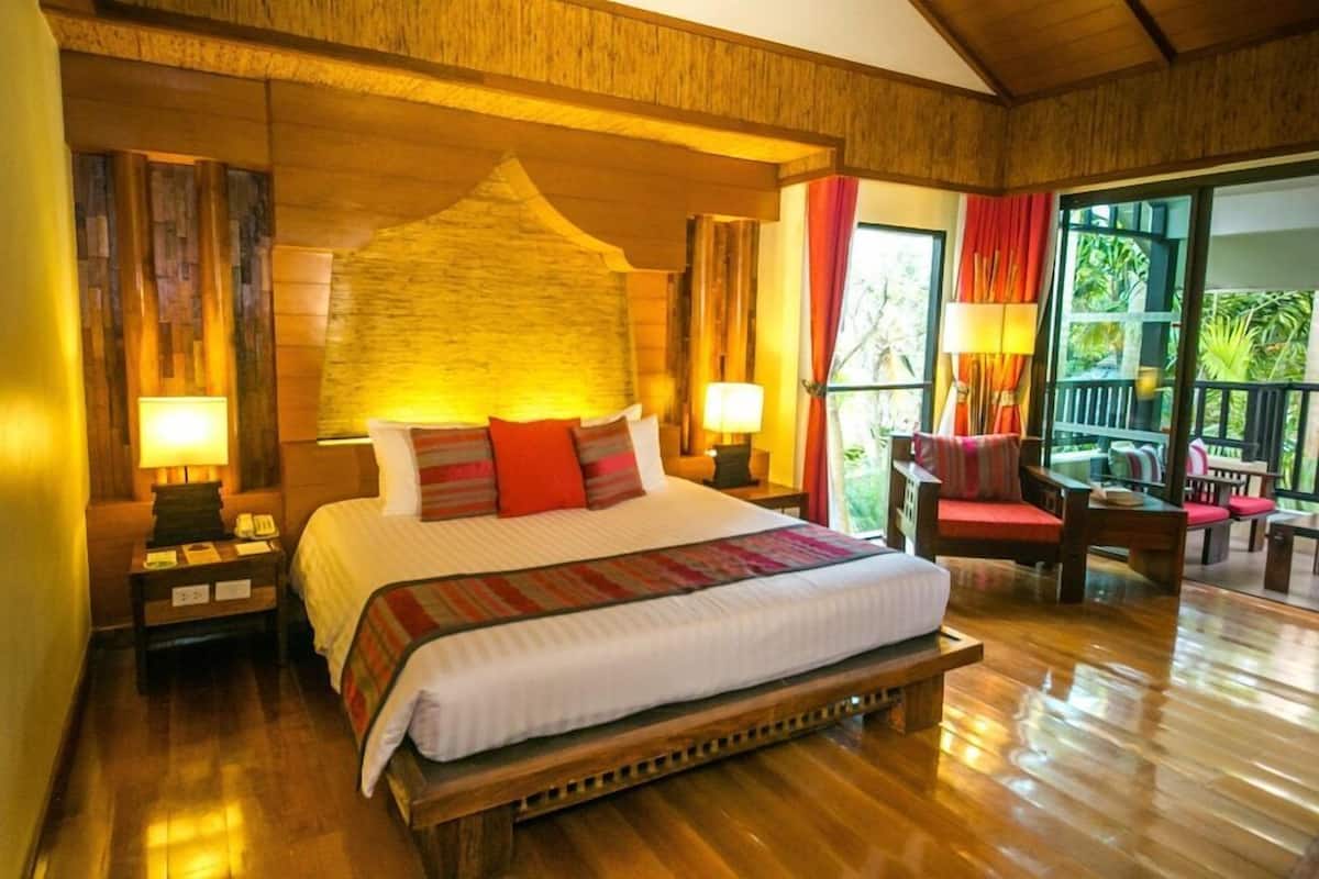 Een groot bed met hemelbed, houten hoofdbord en meerdere kussens. Er is een nachtkastje met een lamp, een houten bankje en een stoel met een rode kussing. De kamer heeft houten vloeren en uitzicht op groen buiten.