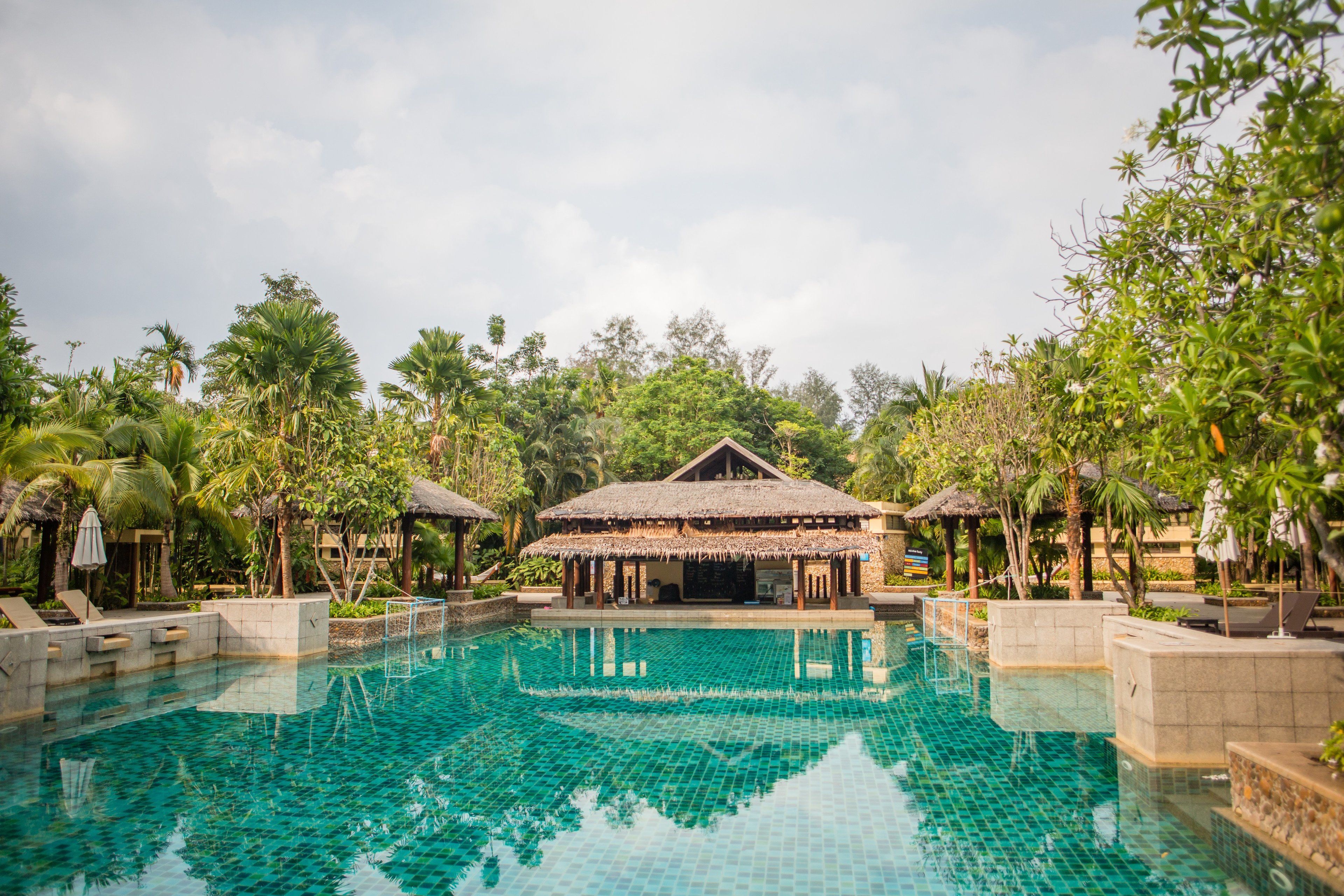 A swimming pool with a thatched-roof structure in the center, surrounded by palm trees and lounge chairs.