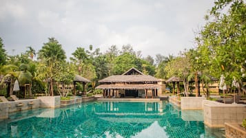 A swimming pool with a thatched-roof structure in the center, surrounded by palm trees and lounge chairs.