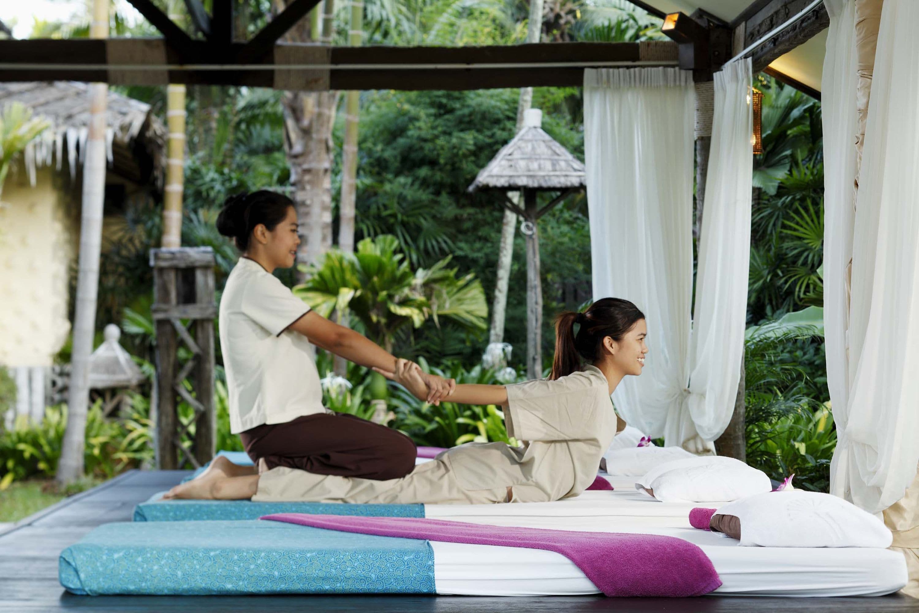 A couple practicing yoga on a mat in a serene outdoor setting with a thatched-roof structure and lush greenery in the background.