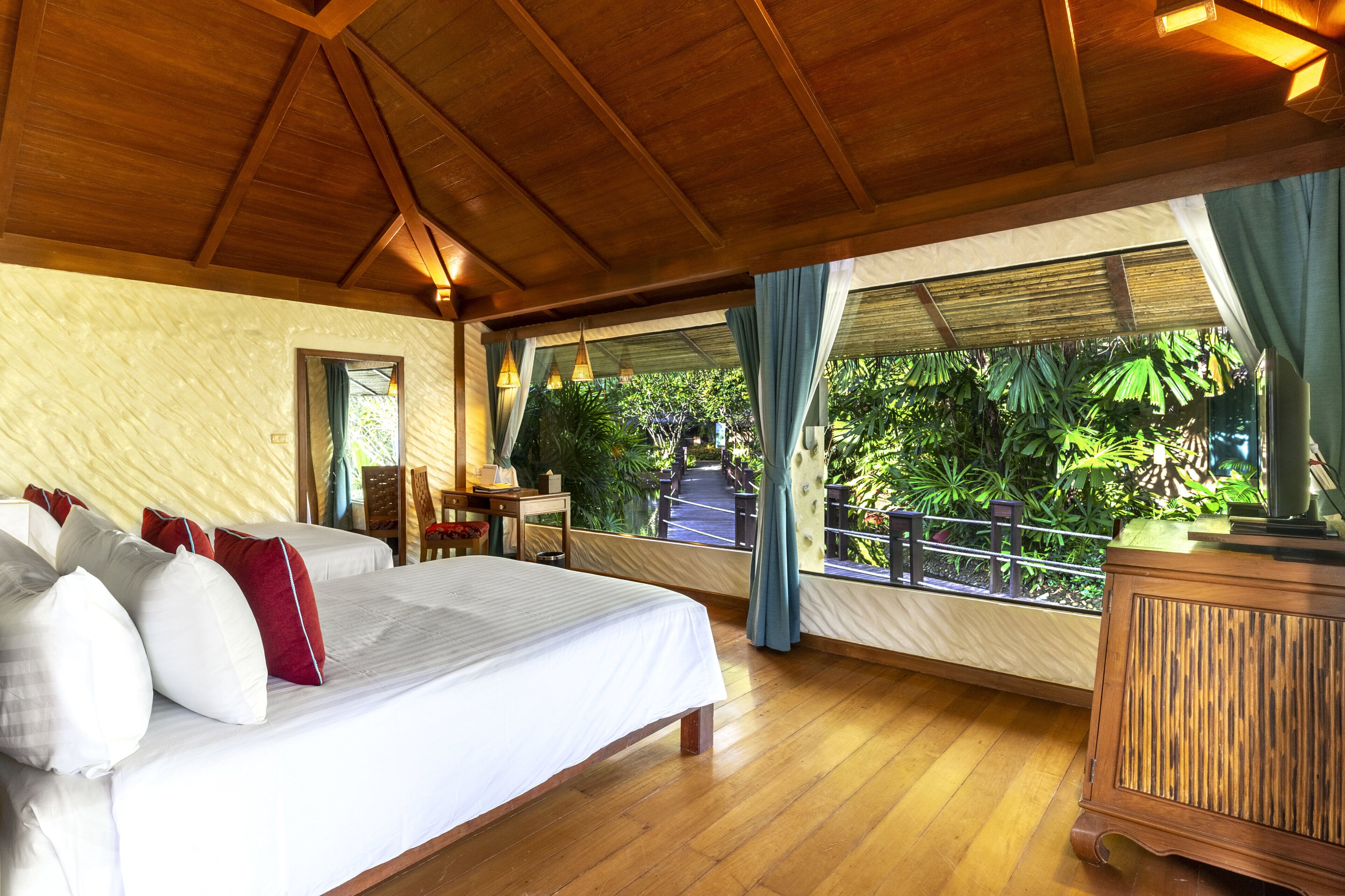 A spacious room with a large bed, wooden ceiling beams, a wooden desk, and a view of lush greenery outside.