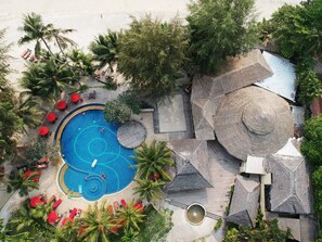 Luchtbeeld van een resort met een centraal gelegen zwembad, omringd door palmbomen en rode parasols.