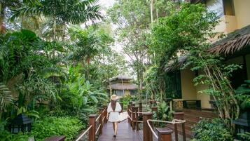 A wooden bridge in a lush garden with a thatched-roof structure in the background.