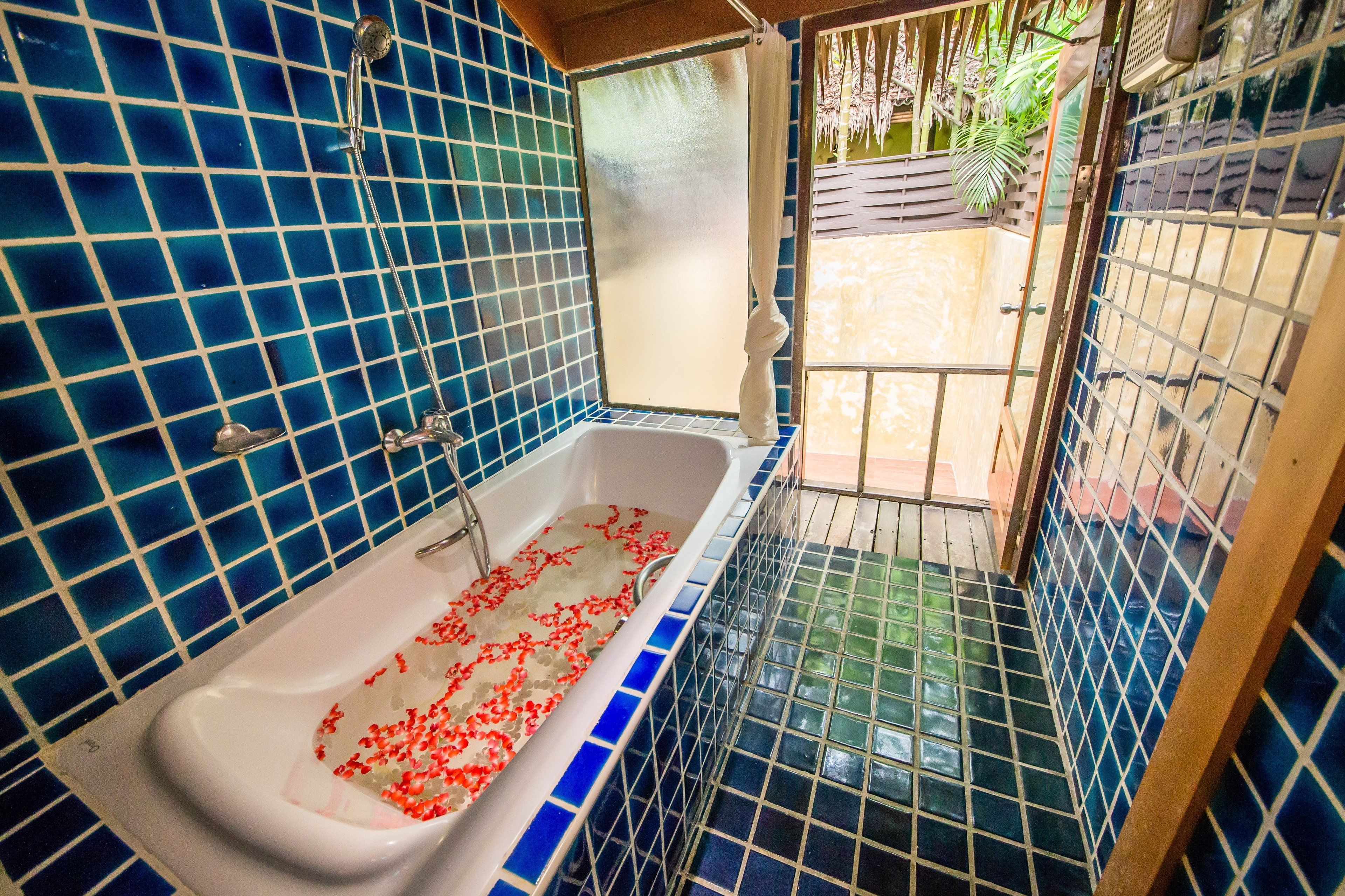 Un baño con paredes de azulejos azules, una bañera blanca con flores rojas y vistas a un jardín a través de una puerta de cristal.