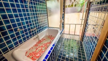 Un baño con paredes de azulejos azules, una bañera blanca con flores rojas y vistas a un jardín a través de una puerta de cristal.