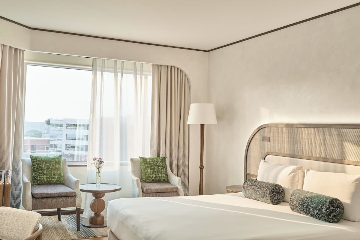 Habitación de hotel con una cama grande, dos sillones, una mesita y una lámpara. La habitación tiene un ventanal con cortinas, y las paredes están pintadas de un color claro.