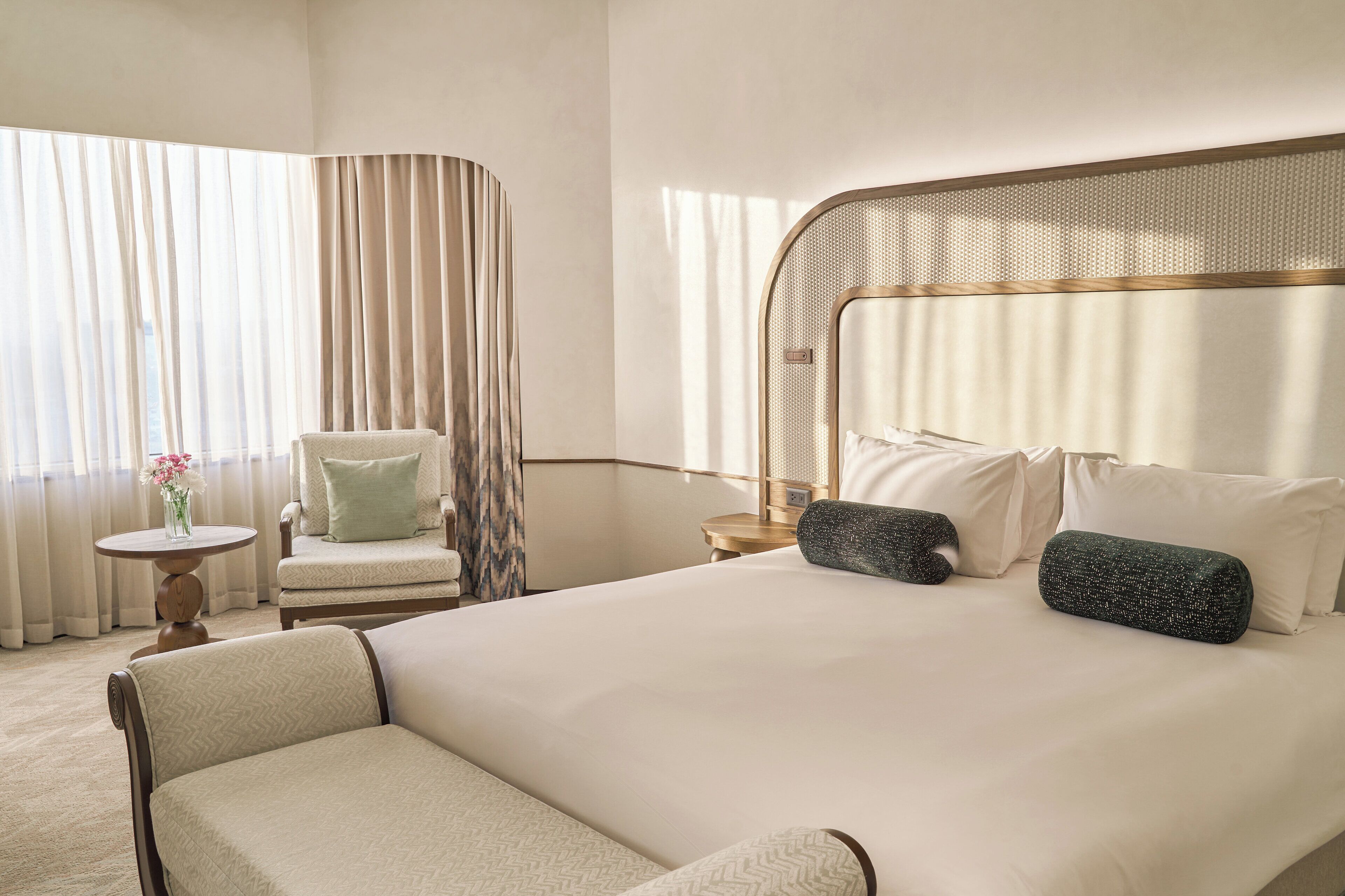 Habitación de hotel con una cama grande, una silla, una mesita con un florero y una ventana con cortinas.