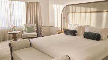Habitación de hotel con una cama grande, una silla, una mesita con un florero y una ventana con cortinas.