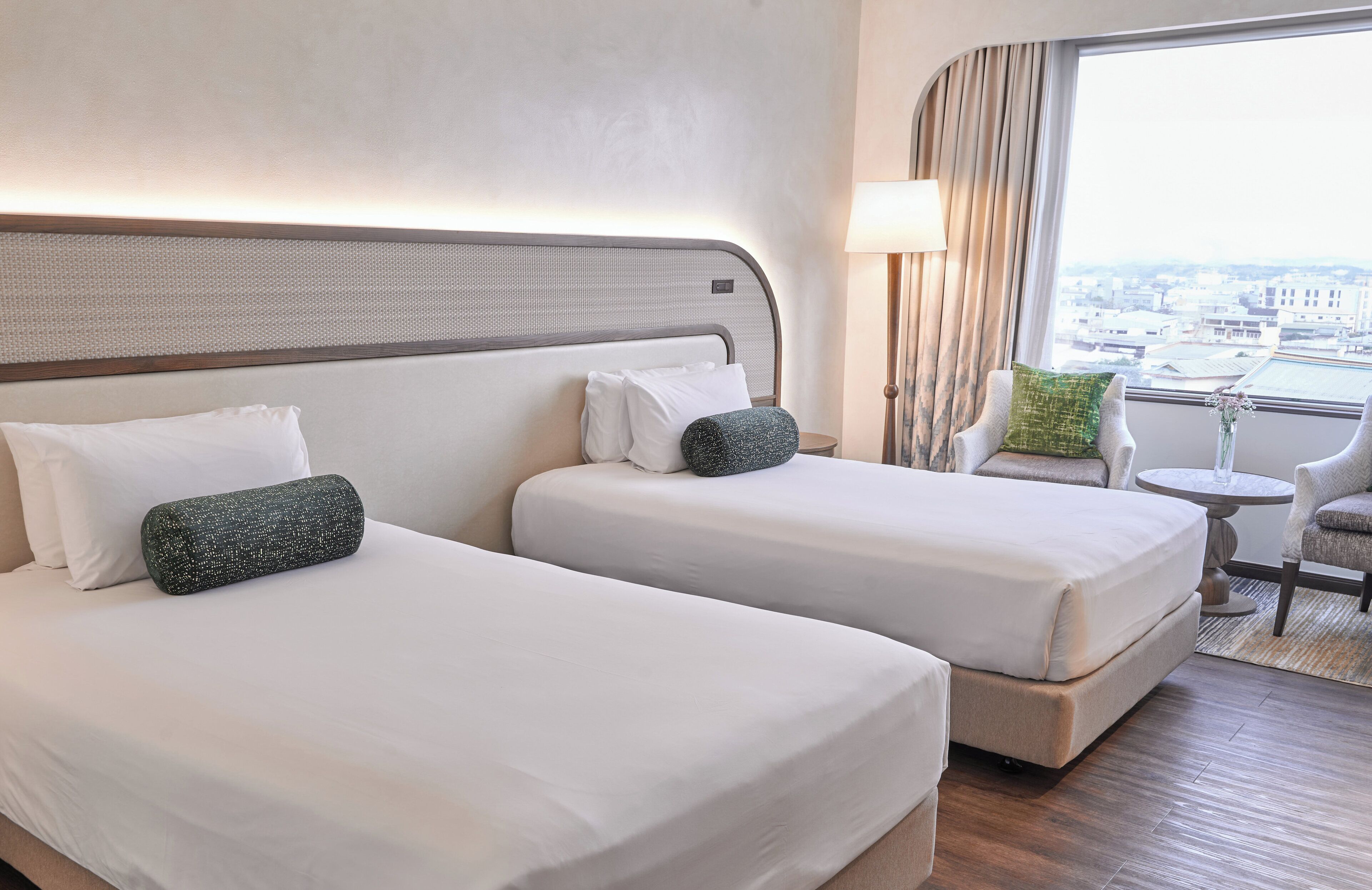 A hotel room with two beds, a small round table, a chair, and a view of the city through a large window.