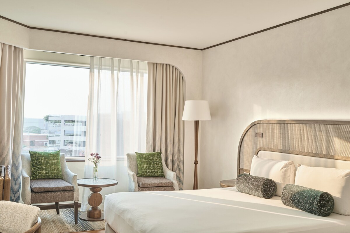 Une chambre d’hôtel avec un lit, deux fauteuils, une petite table, une lampe et une vue sur les immeubles à travers de grandes fenêtres.