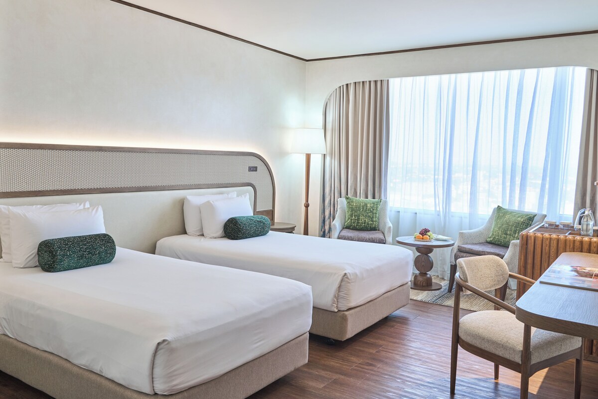 Une chambre d’hôtel avec deux lits, une chaise, une table et une vue sur la ville grâce à de grandes fenêtres.