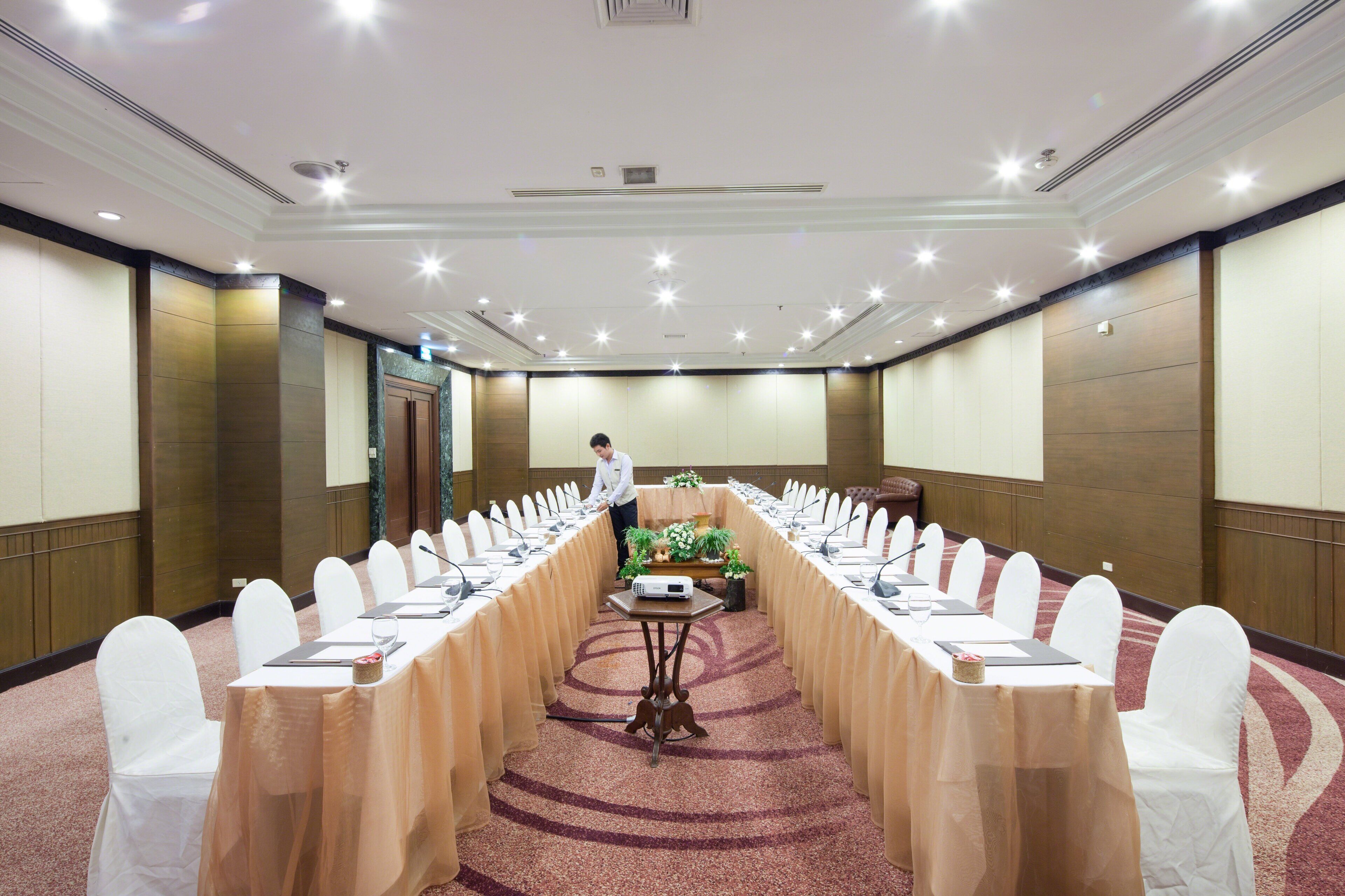 A conference room with a long table, white chairs, and a person standing at the front.