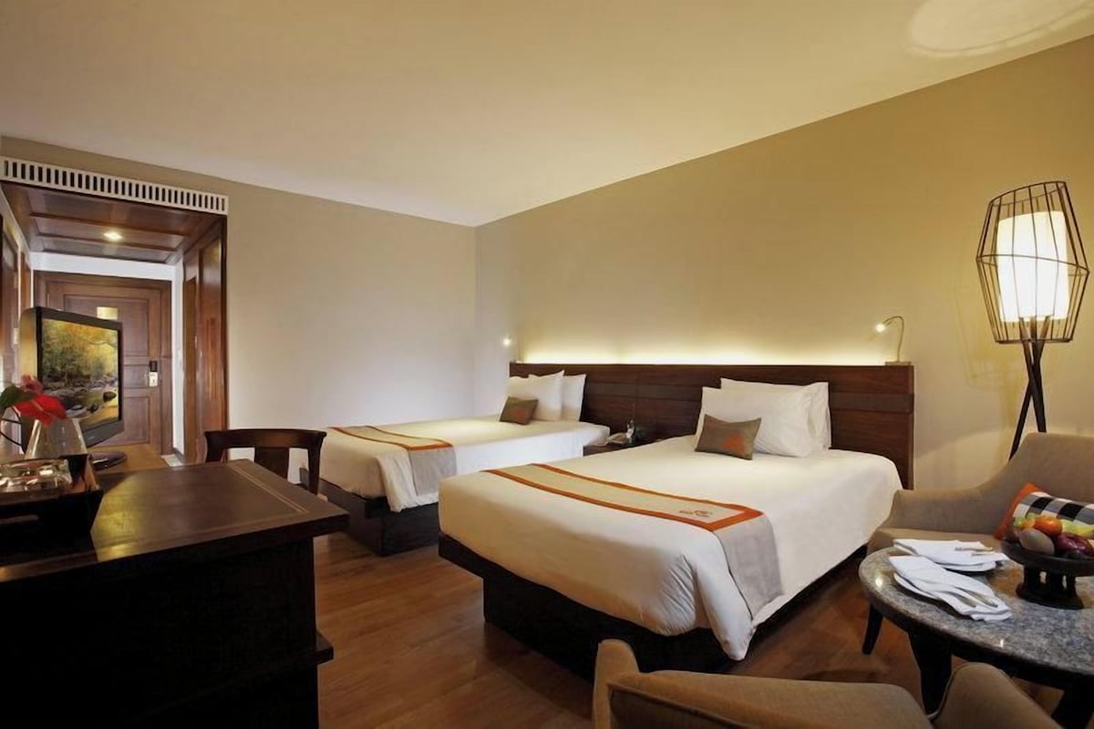 A hotel room with two beds, a desk, a chair, and a lamp.