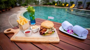 A wooden table with a meal consisting of a burger, fries, and a drink, set against a poolside view.