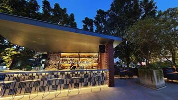 An outdoor bar area with a tiled backsplash, a person behind the bar, and a seating area with chairs and a table.