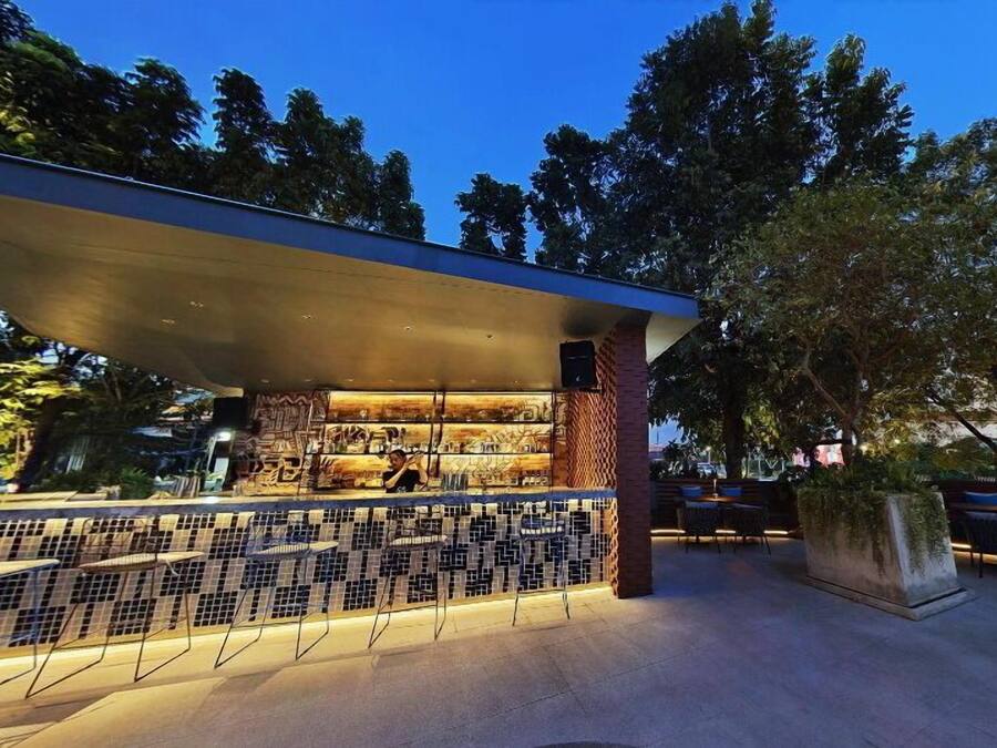 An outdoor bar area with a tiled backsplash, a person behind the bar, and a seating area with chairs and a table.