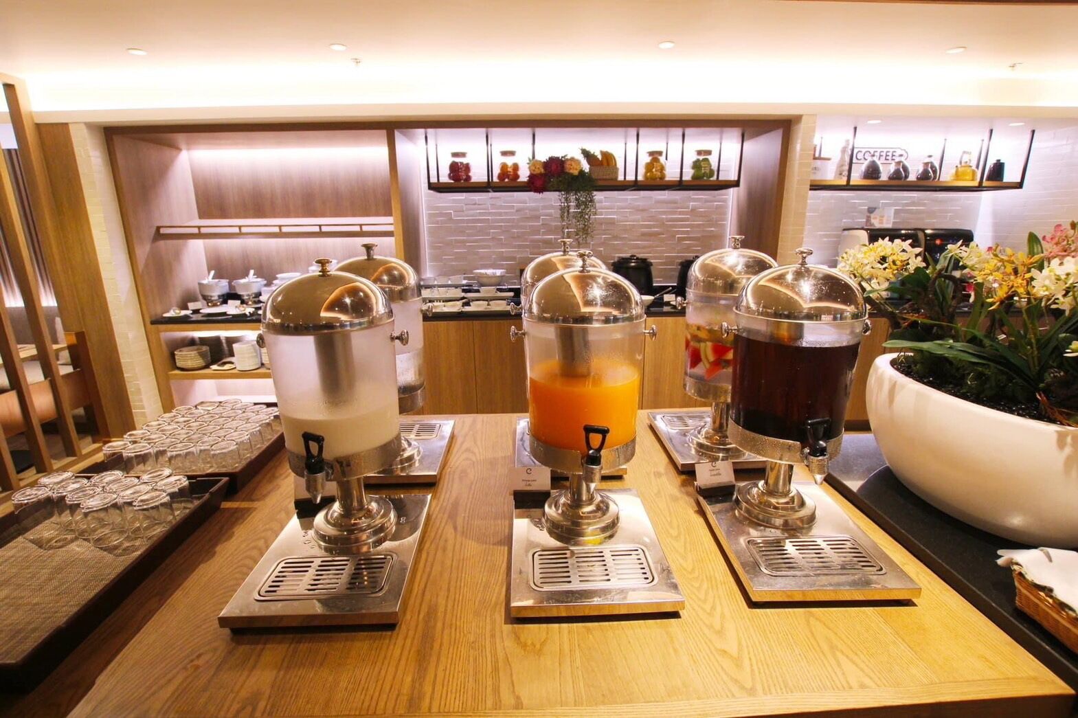 A breakfast bar with three beverage dispensers, a coffee machine, and a large flower pot.