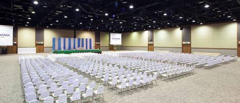 A large conference room with rows of white chairs, a stage with a blue backdrop, and a projection screen.