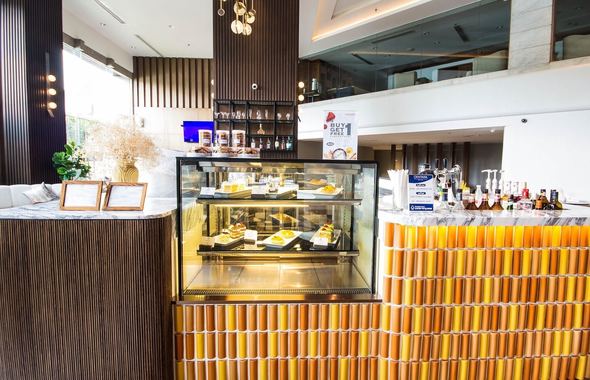 A modern bar area with a glass display case of desserts, a counter with a sign offering a free item, and a variety of bottles and glasses on the counter.