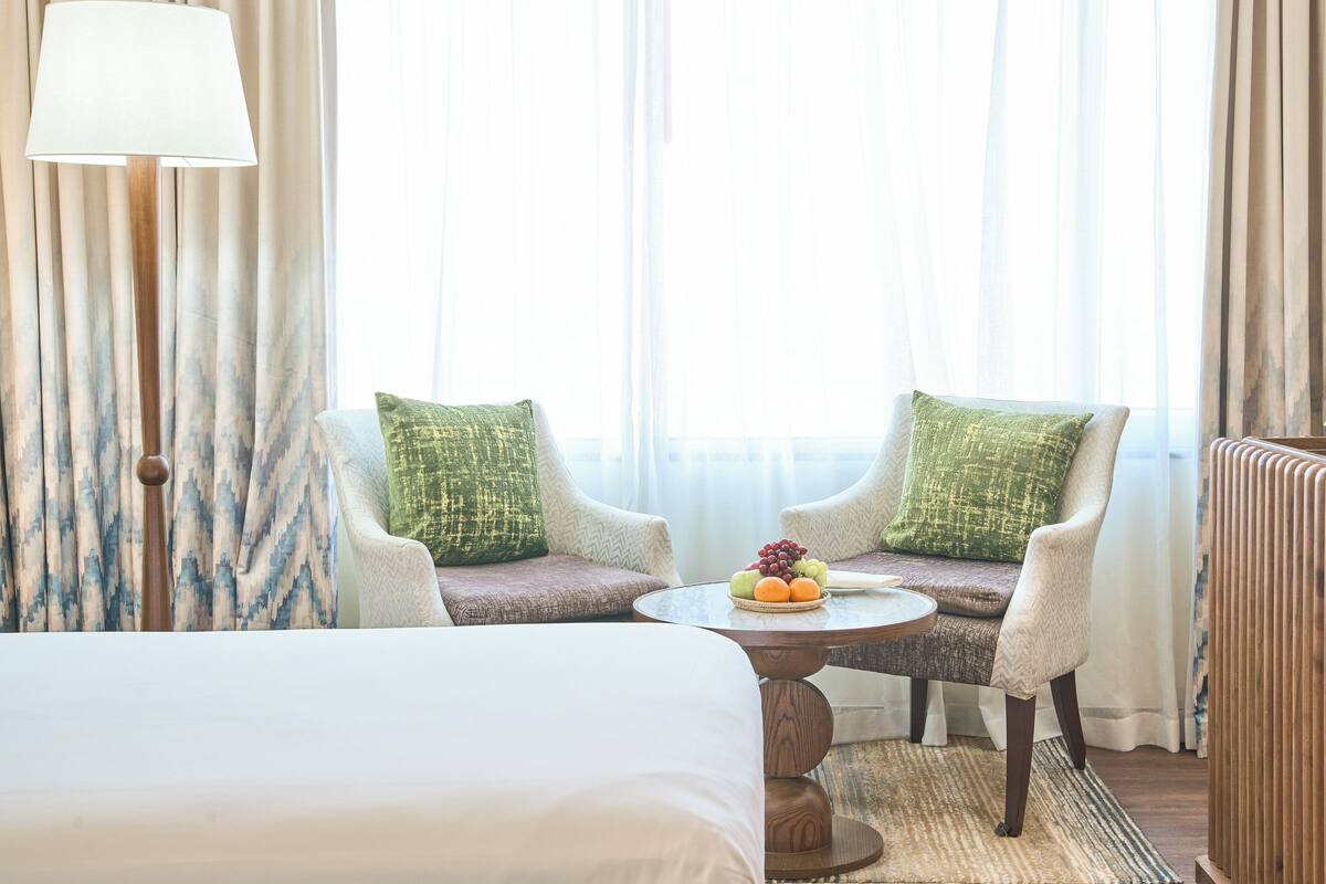 Una habitación de hotel con una cama, dos sillones, una mesita redonda con fruta y una ventana con cortinas.