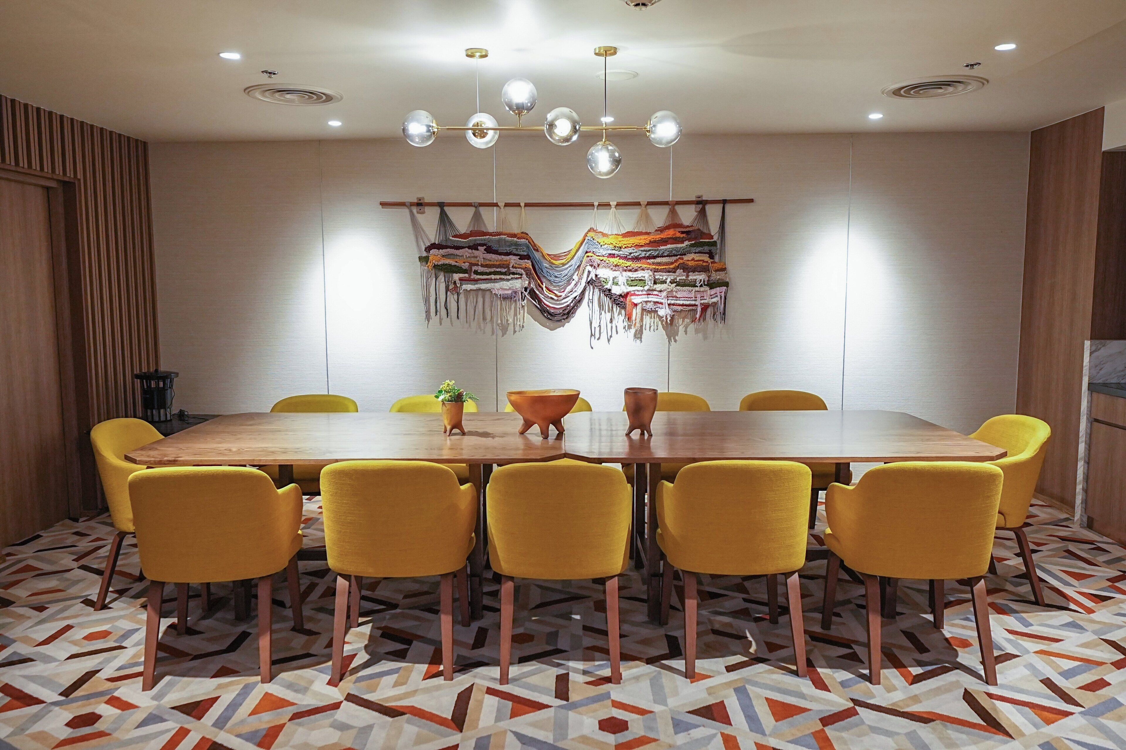 Een vergaderruimte met een houten tafel, gele stoelen, een kamer met een patroon vloer en een hangend textielkunstwerk.